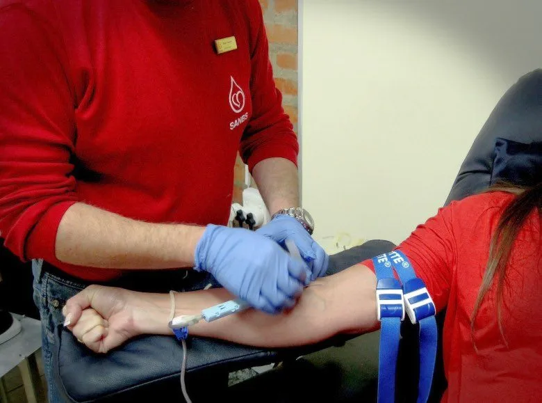 Infirmier en blouse rouge SANG avec gants bleus posant un garrot et l'aiguille de prélèvement sur le bras d'une donneuse en centre de collecte