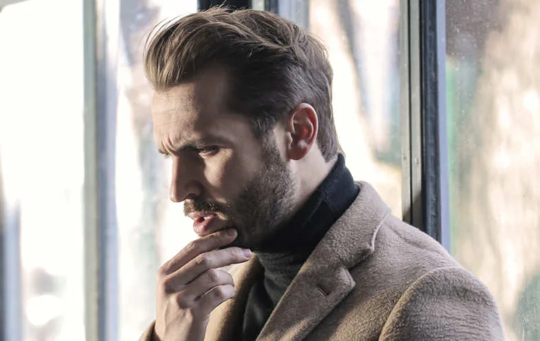 Portrait en profil d'un homme barbu pensif vêtu d'un manteau beige et d'un col roulé sombre devant une fenêtre lumineuse