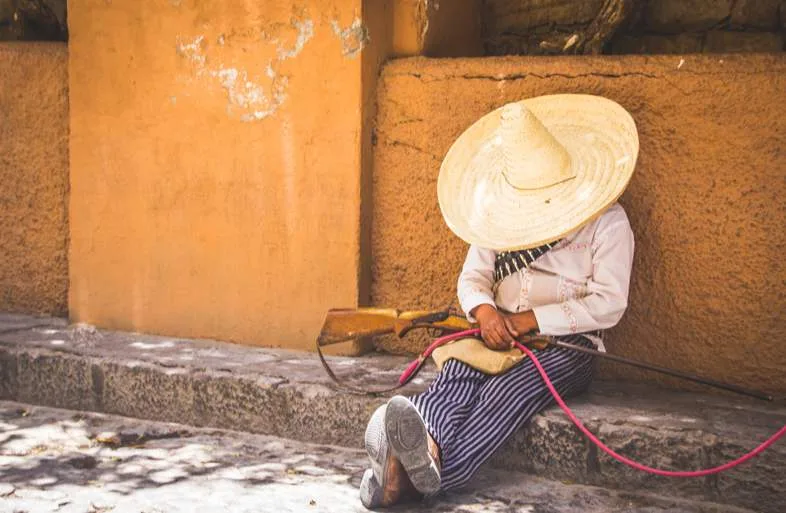 Personnage en tenue traditionnelle mexicaine assis contre un mur ocre avec sombrero et carabine, scène évoquant le contexte rural historique mexicain
