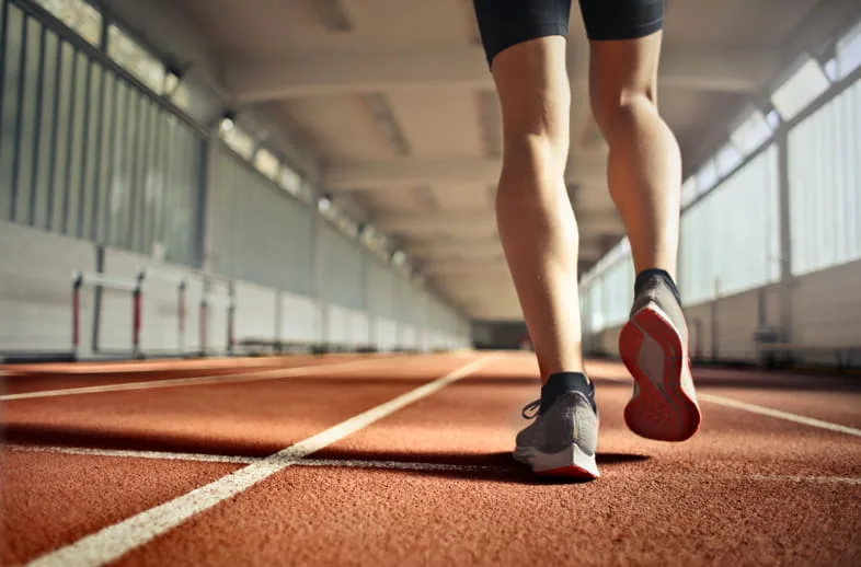 Vue rapprochée de jambes et chaussures de course d'un coureur en mouvement sur piste d'athlétisme indoor avec haies en arrière-plan