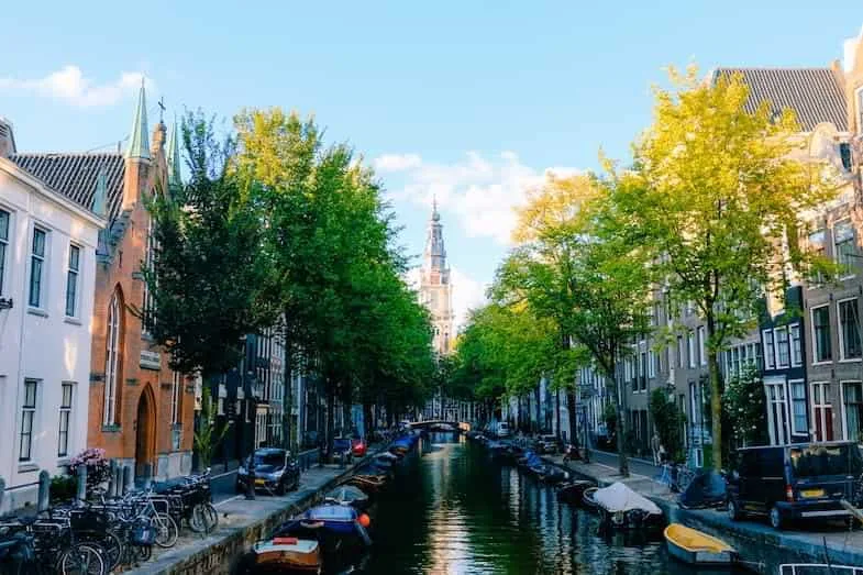 Vue ensoleillée d'un canal d'Amsterdam bordé d'arbres et d'immeubles historiques avec église en arrière-plan, paysage typique du centre néerlandais