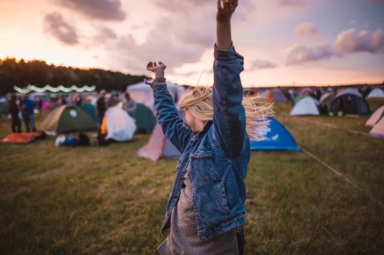 Jeune femme aux cheveux blonds en veste en jean lève les bras au coucher du soleil dans un campement de festival contemporain héritier de l'esprit hippie