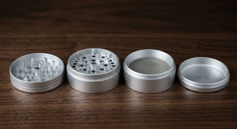 Grinder weed en quatre parties en aluminium argenté entièrement démonté et aligné sur une surface en bois foncé, chambre à kief ouverte exposant la grille tamis