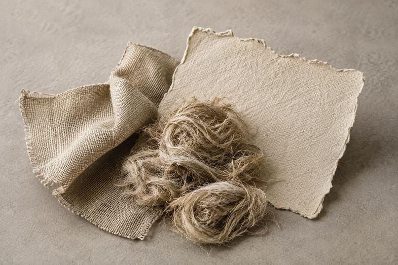 Fibre de chanvre brute, toile tissée et papier artisanal Fibre de chanvre brute posée entre une toile tissée écrue repliée et une feuille de papier artisanal, sur un fond de pierre grise